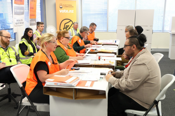 People are sitting at tables. People in orange vests are practicing issuing votes to other people. There are people in yellow vests observing. 
