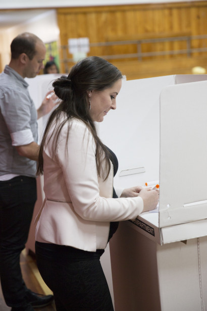 A woman in a voting booth on election day, filling in her voting paper. In the background, a man is in another voting booth, filling in his paper.